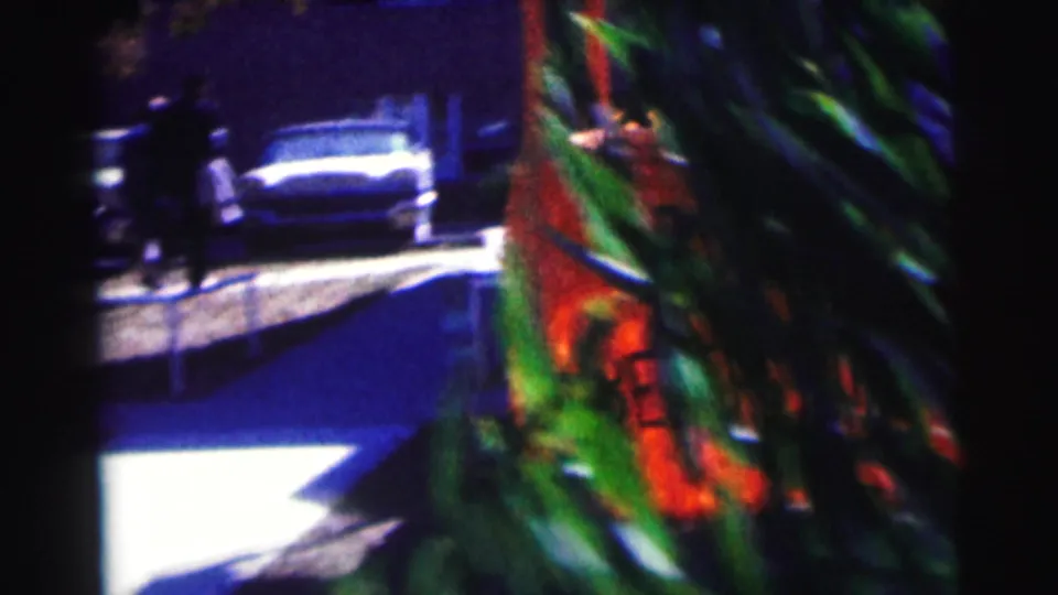 A 1963 Bowling Green University street-side view shows a parked car, distant people and a partially hidden root beer sign in saturated 8mm color.
