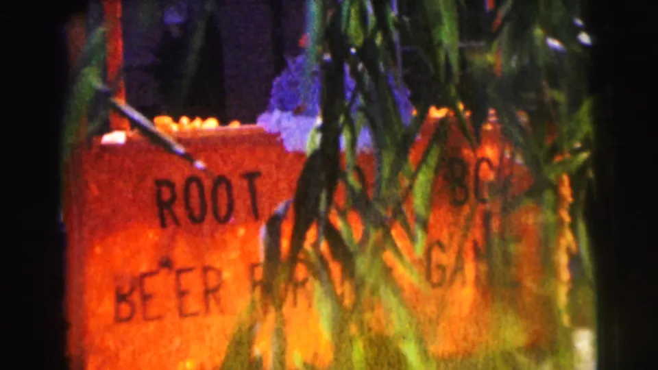 Close 1963 home-movie view of the orange root beer sign, with handmade lettering and hanging greenery obscuring part of the message.