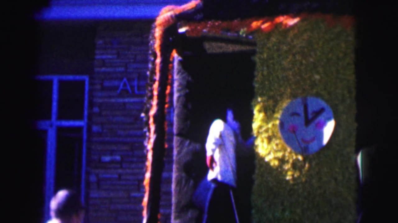 Nighttime 8mm frame at Bowling Green University showing a decorated doorway with a green panel, round face cutout, and a person in white emerging.