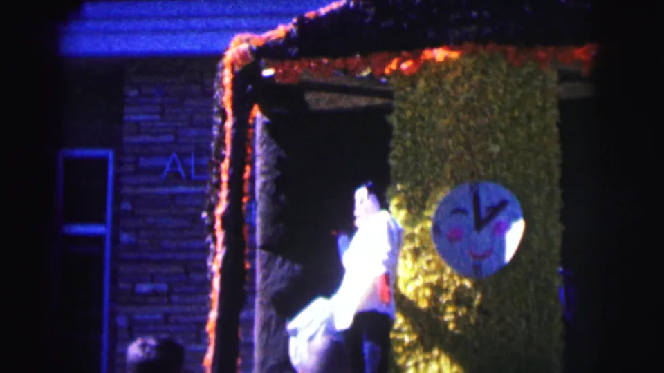 A person in white stands near the decorated house entrance, with green covering, orange trim, and a round face decoration visible.
