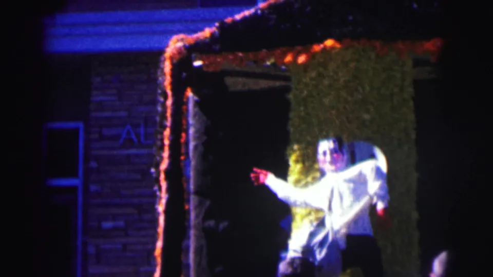 Nighttime Bowling Green University frame showing a person gesturing from a decorated doorway with garland, green covering, and a round face cutout.