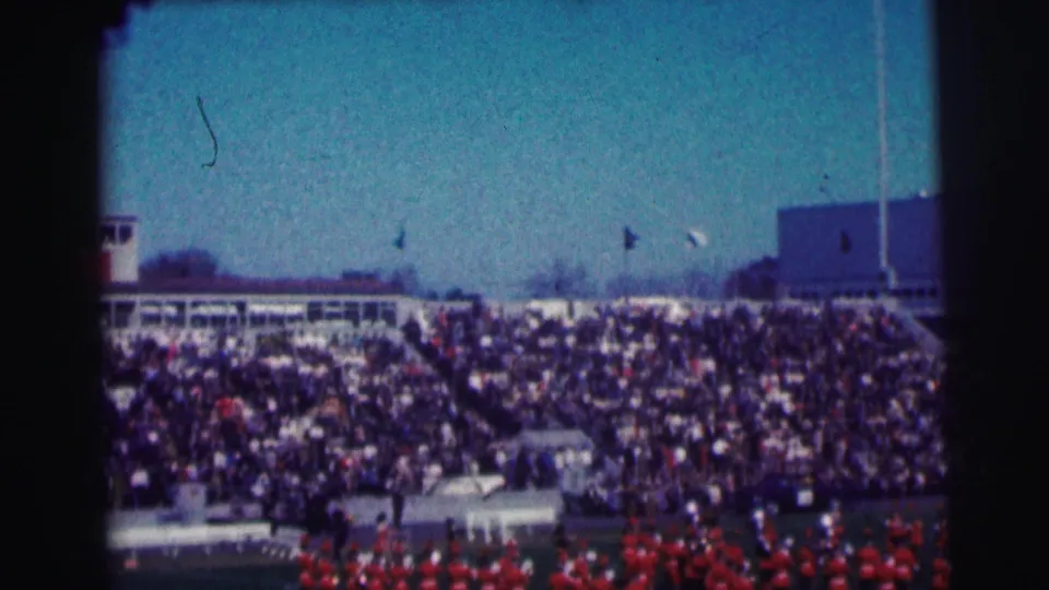 Handheld 8mm view of a packed Bowling Green University stadium, with flags above the stands and band members in red below.