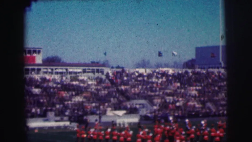 Distant 1963 campus stadium shot showing packed stands, flags, and a red-uniformed marching band near the field edge.