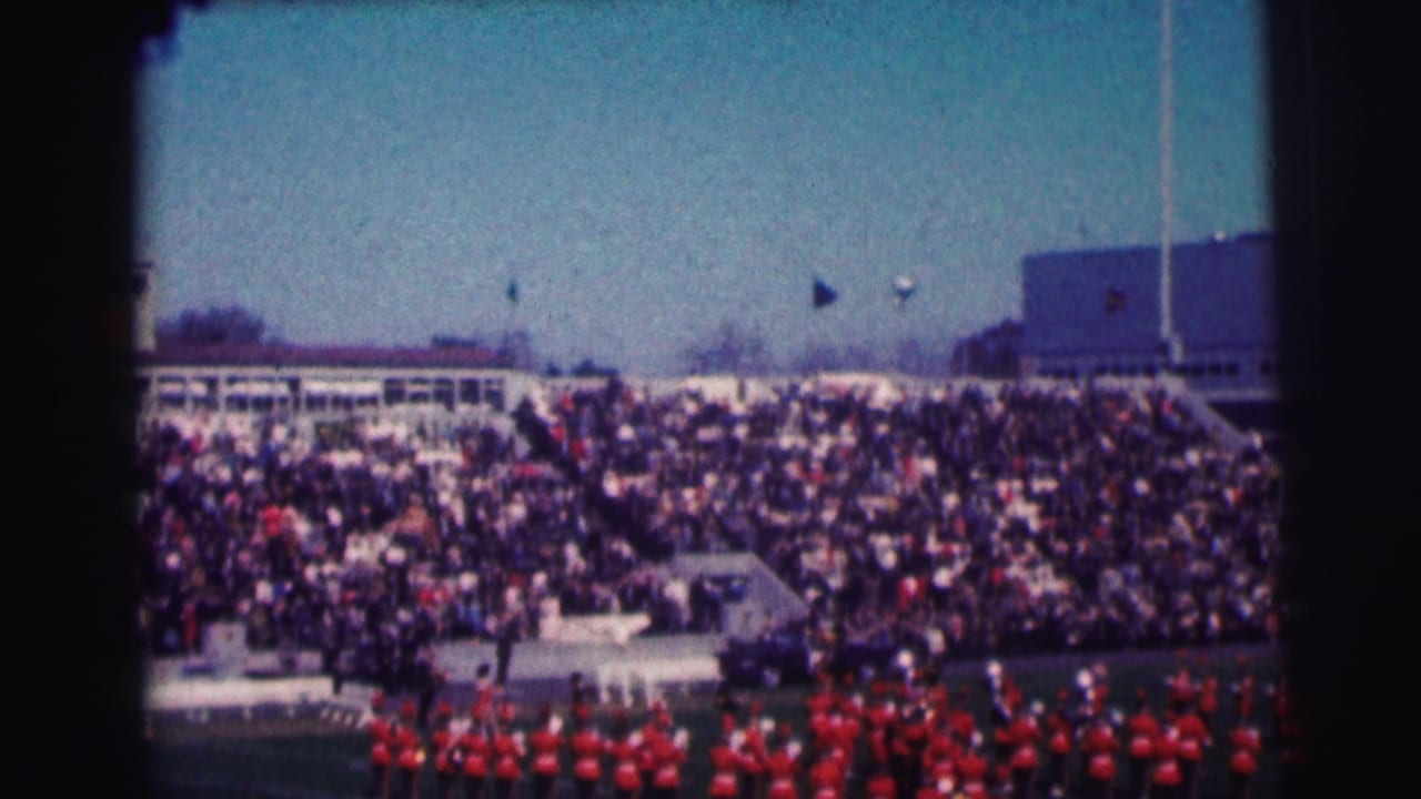 1963 8mm view of crowded Bowling Green University stadium bleachers with a marching band in red uniforms on the field.