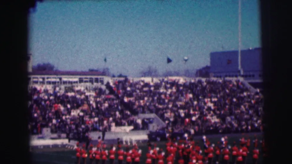 1963 8mm view of crowded Bowling Green University stadium bleachers with a marching band in red uniforms on the field.