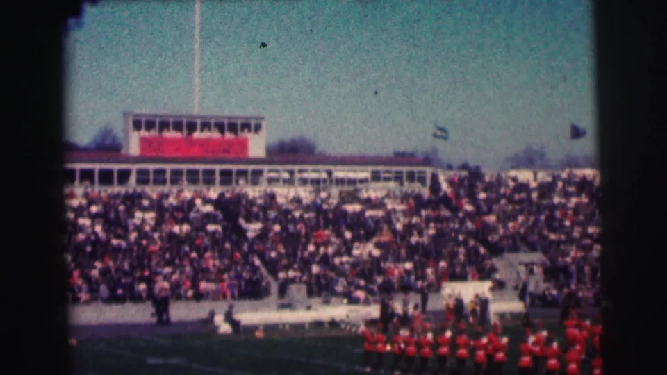 Wide 1963 8mm stadium view of Bowling Green University crowds, press box, flags, and red-uniformed marching band.