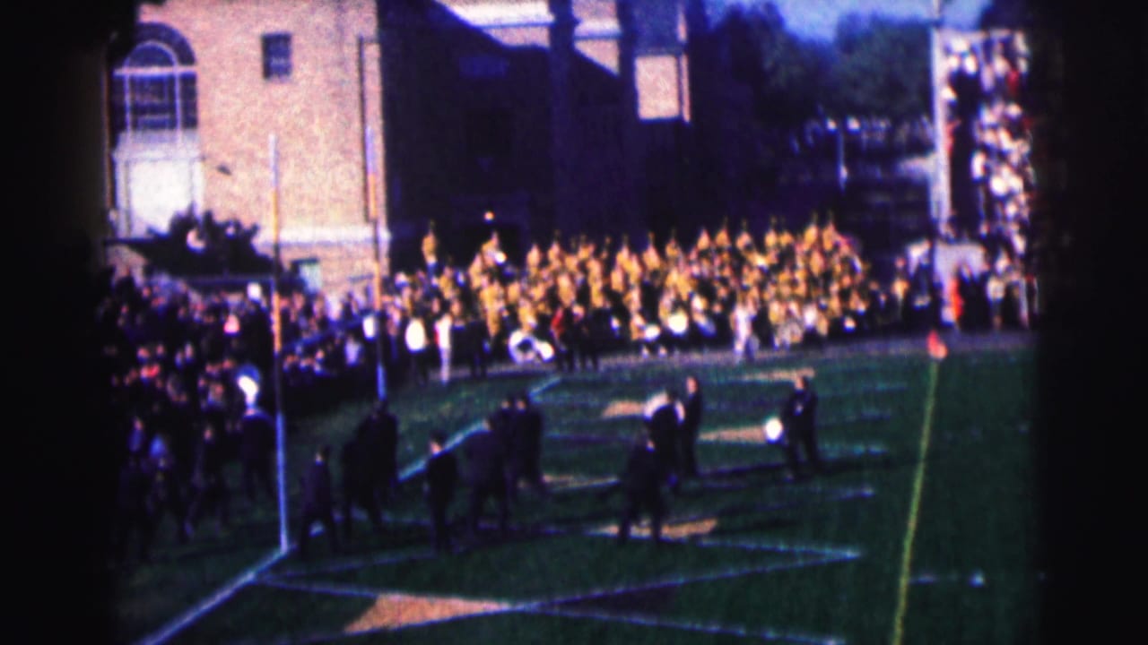 1963 Bowling Green University home-movie frame showing a marching band on a football field before spectators and brick campus buildings.