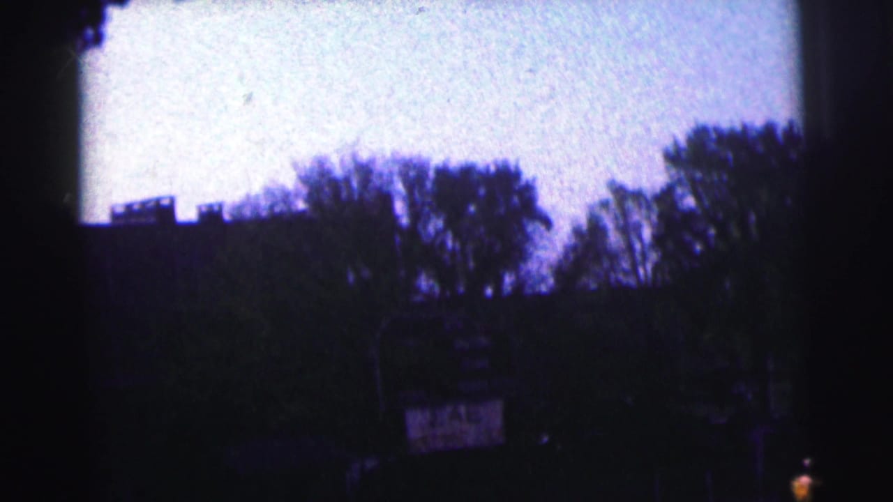 Dark outdoor scene with tree silhouettes, rooflines, and a pale sky.