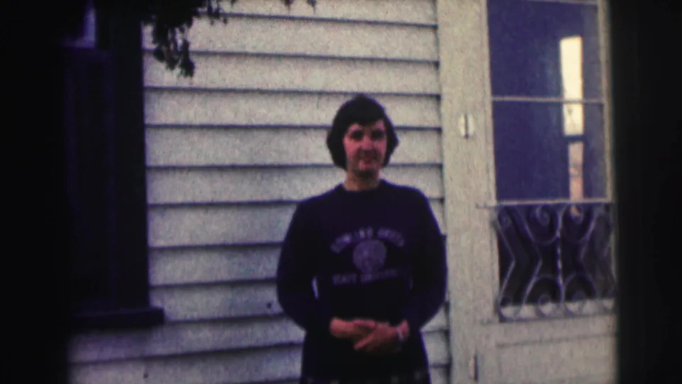 Woman with short dark hair stands outside a light-sided building in 1963 Bowling Green University home-movie footage, with lettering visible on her sweatshirt.