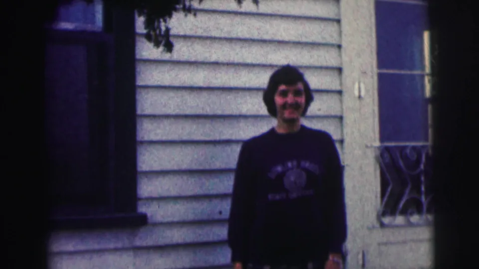 The final frame holds on the woman in her university sweatshirt outside a light-sided house in 1963 home-movie footage.