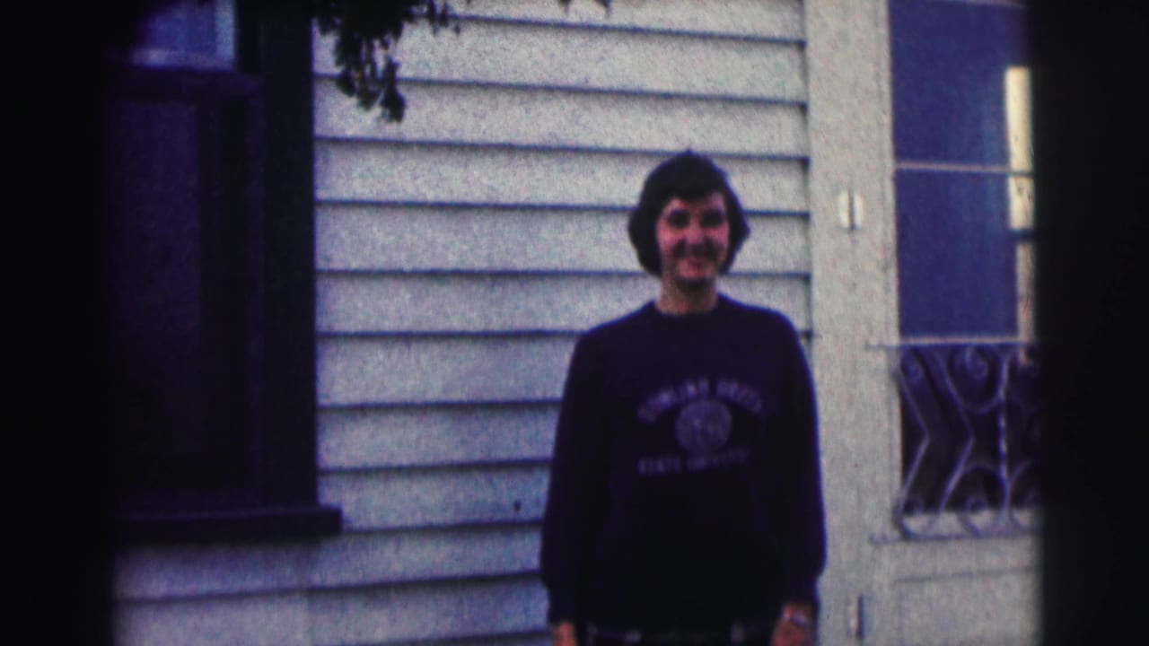 Young man in a Bowling Green State University sweatshirt stands smiling outside a light-sided house in 1963 home-movie footage.