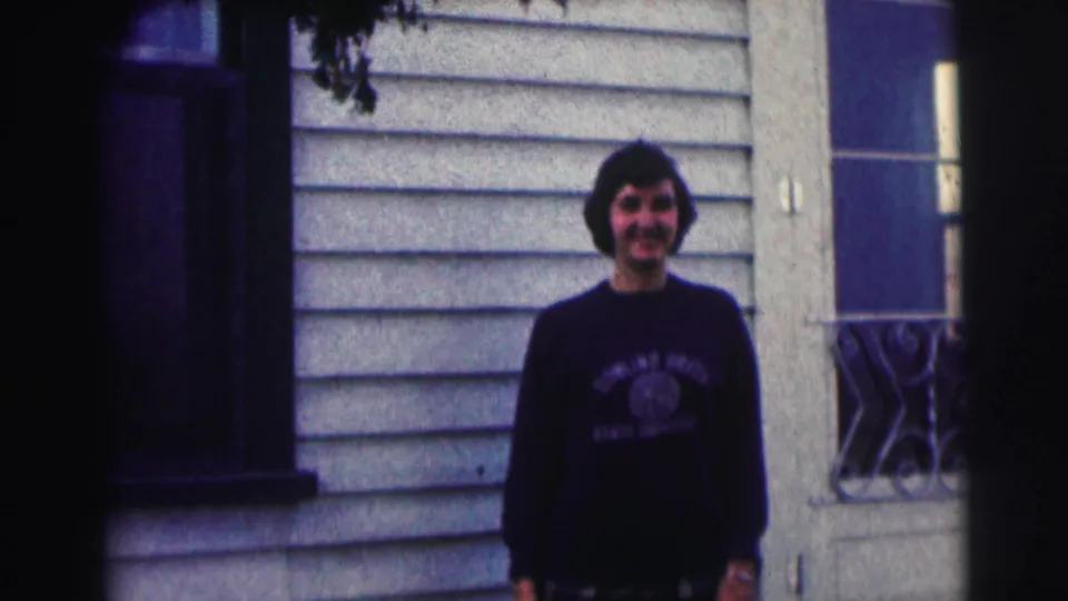 Young man in a Bowling Green State University sweatshirt stands smiling outside a light-sided house in 1963 home-movie footage.