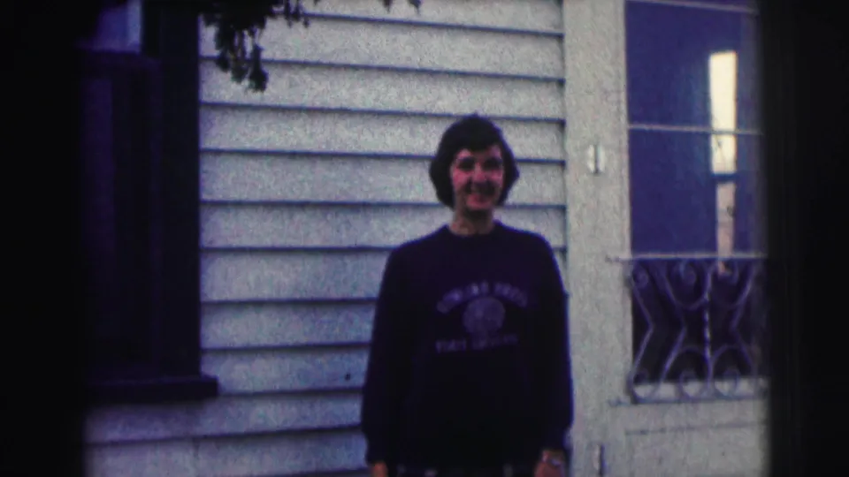 Young man in a dark Bowling Green State University sweatshirt stands centered outside, framed by a window and light house siding.