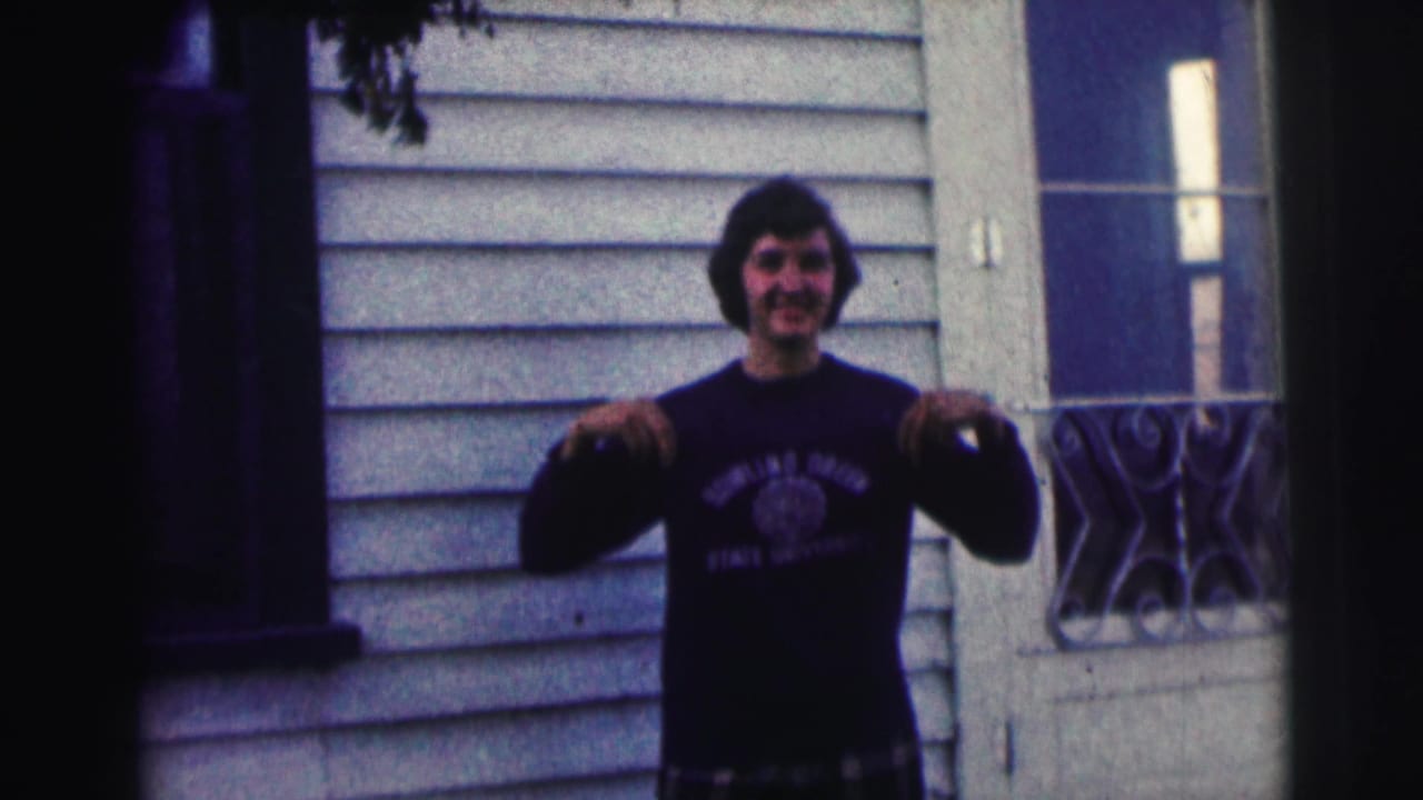 Young man in a Bowling Green State University sweatshirt stands by a white-sided house in 1963 home-movie footage, smiling toward the camera.