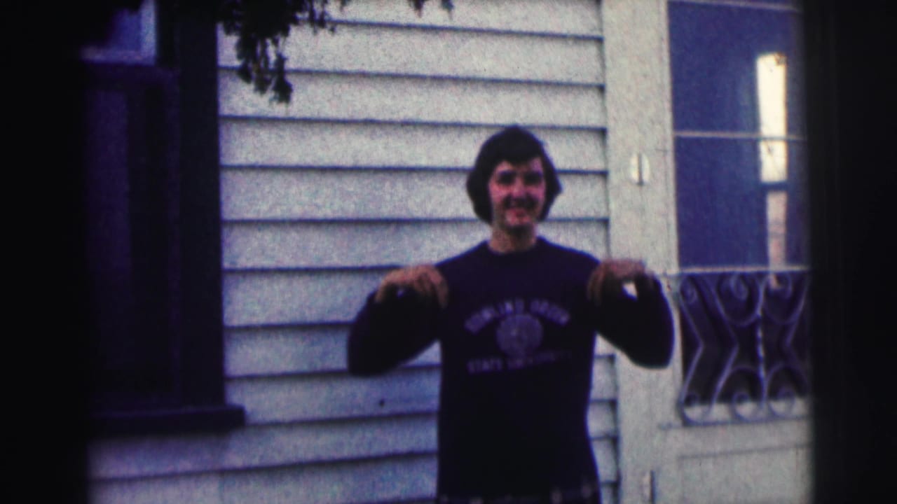 Smiling man in a dark sweatshirt gestures in front of house siding, with a door and railing nearby.