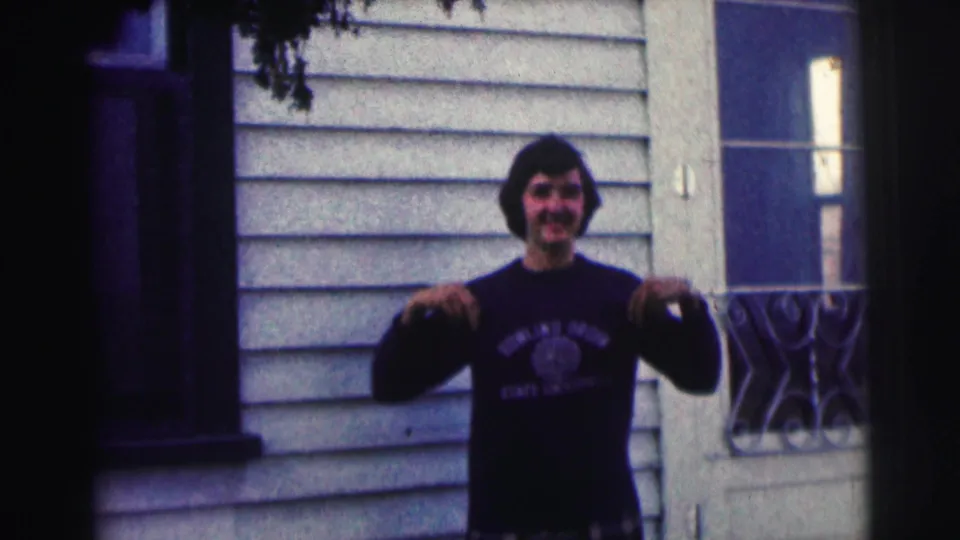 Smiling young man gestures at his Bowling Green State University sweatshirt near a door and decorative railing in 1963 home-movie footage.