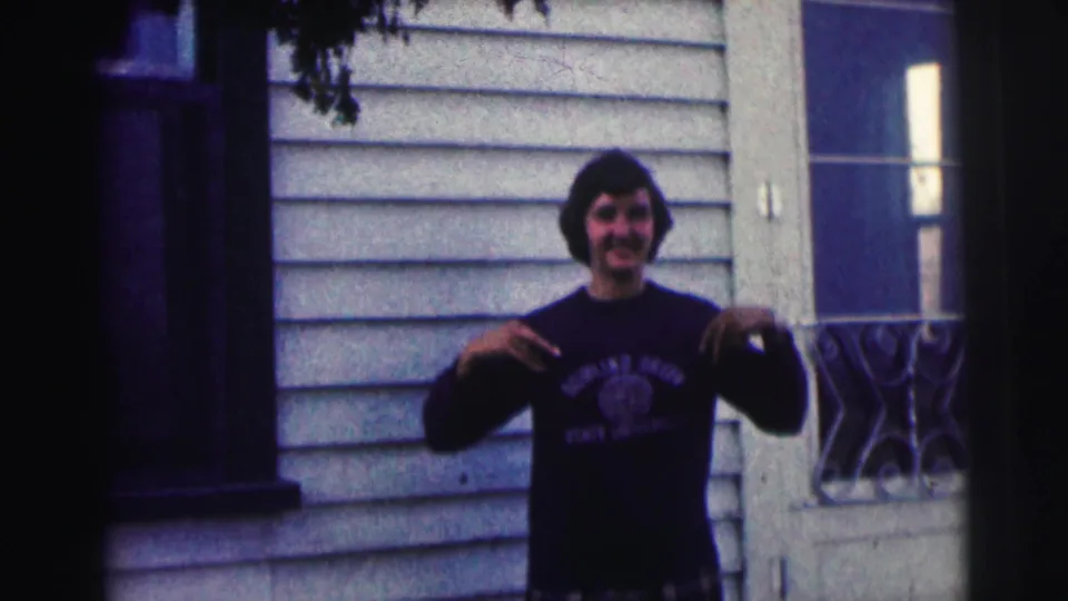 Young man in a dark Bowling Green sweatshirt stands outside a white-sided residence, one hand lowered and the other near his chest.