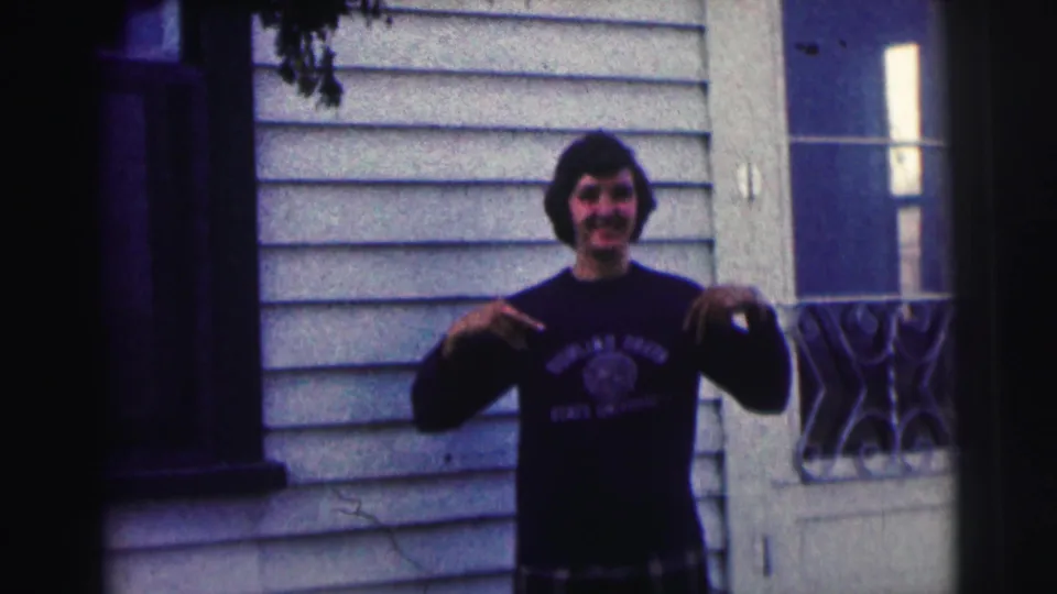 Young man in a Bowling Green State University sweatshirt points at his chest while standing outside a white-sided house in the 1963 clip.