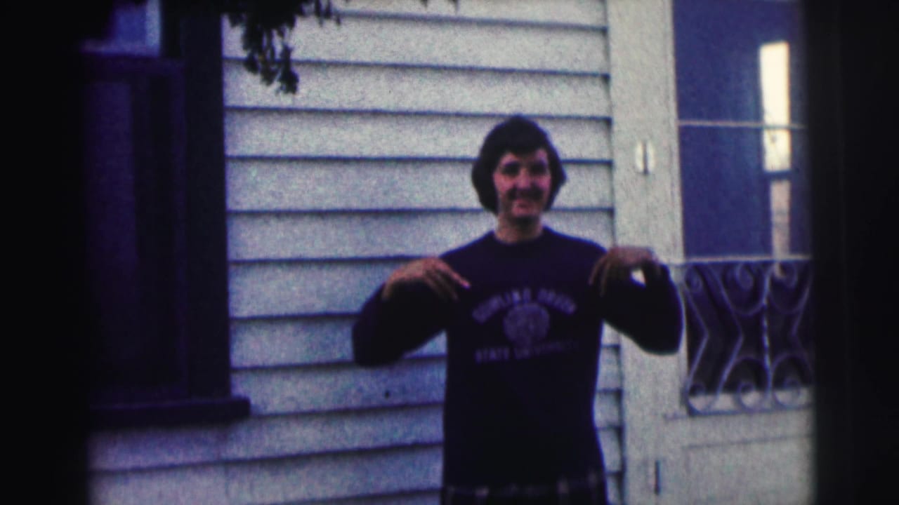 Young man outside a sided house points toward the lettering on his dark shirt.