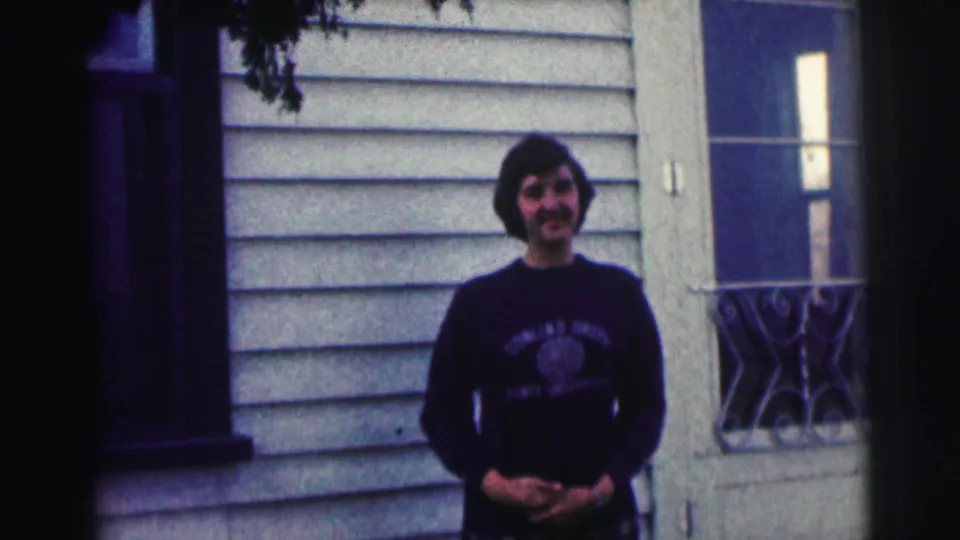 A young man in a Bowling Green shirt stands outside a light-sided house, hands clasped and facing the camera.
