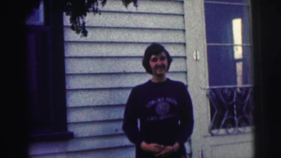 The 1963 home-movie frame shows the man smiling in his Bowling Green shirt beside a door and window.