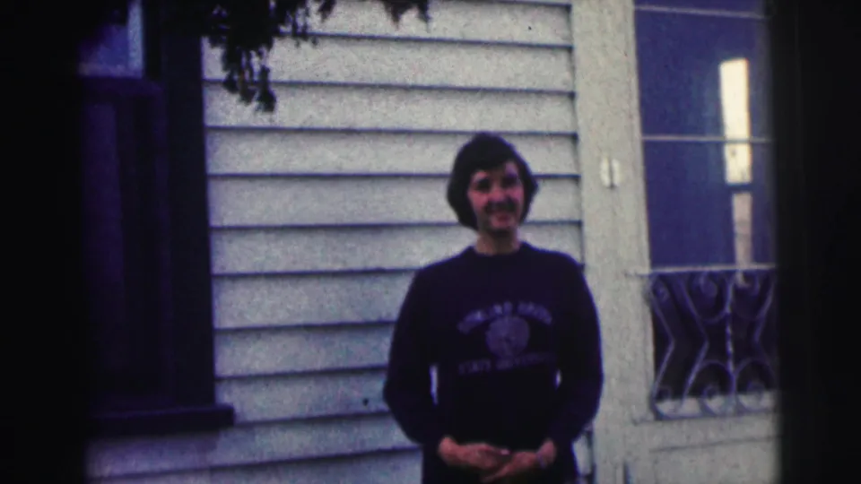 Young woman in a Bowling Green State University sweatshirt smiles outside a light-sided house in 1963 home-movie footage.