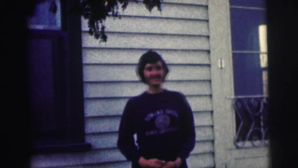 Soft 8mm frame of a young woman in a university sweatshirt standing before light siding and dark trim.