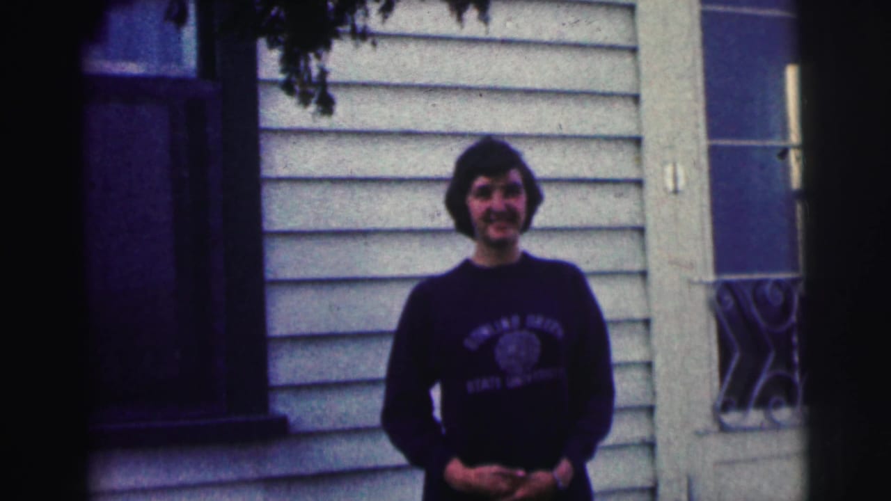 Young woman in a Bowling Green State University sweatshirt smiles outside a light-colored house in 1963 home-movie footage.