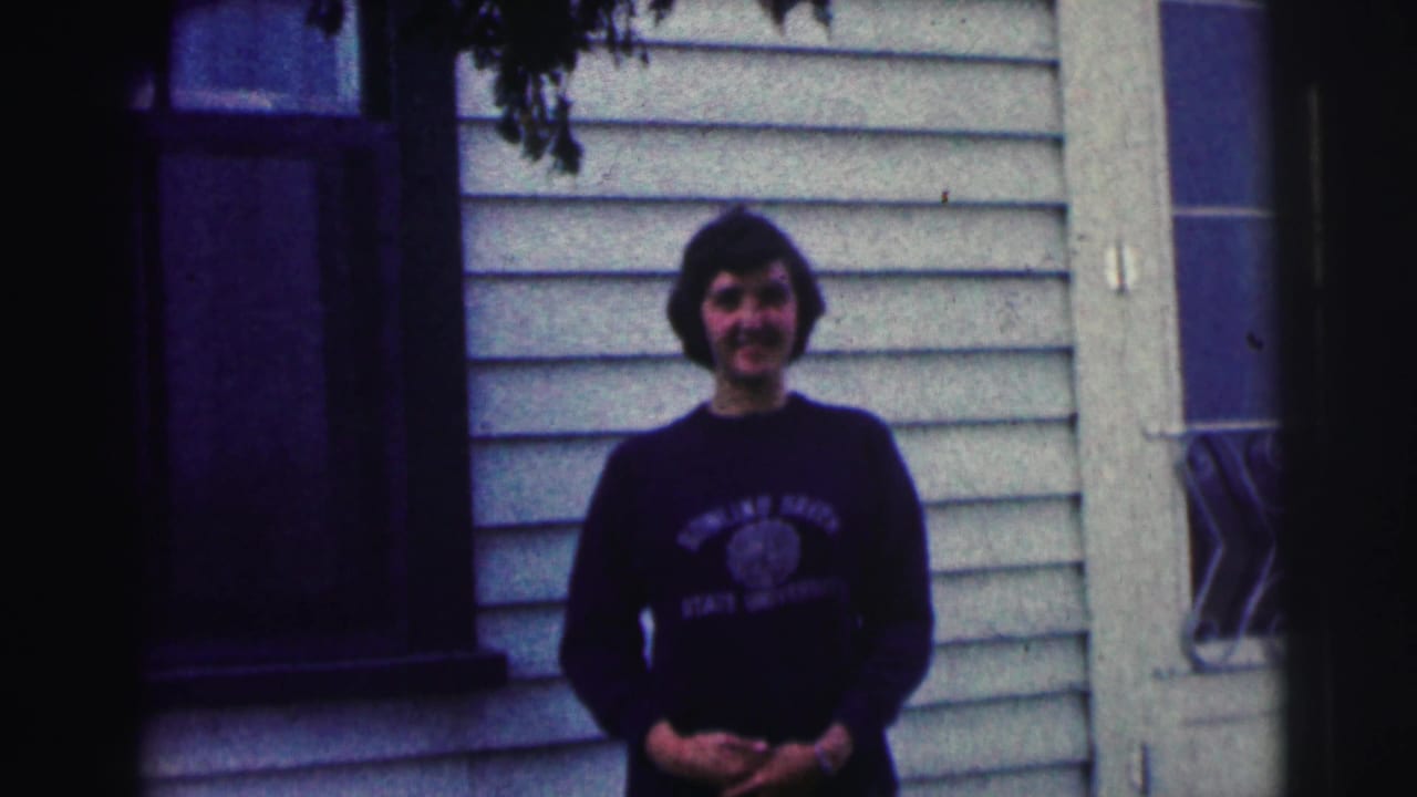Smiling young woman in a Bowling Green State University sweatshirt stands with hands clasped outside a light-sided house.