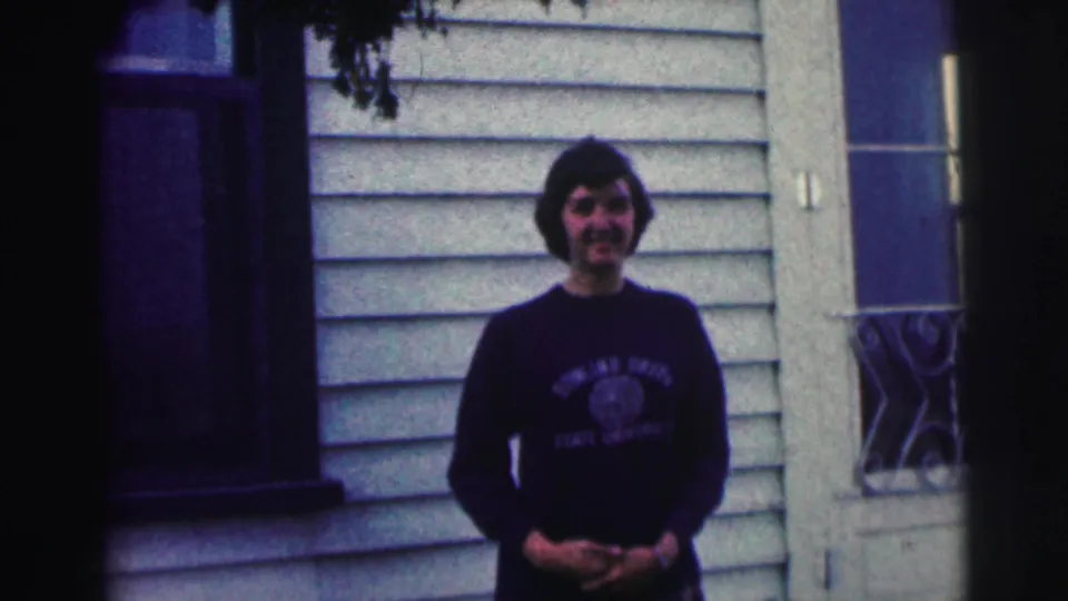 In the 1963 8mm frame, a young woman in collegiate clothing faces the camera outside a light-sided house.