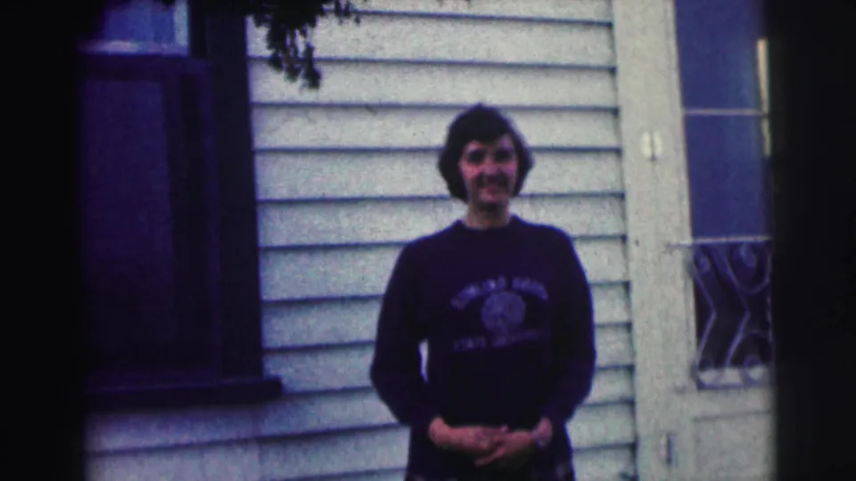 Young woman in a Bowling Green State University sweatshirt smiles outside a house, framed by siding, window, railing, and branches.