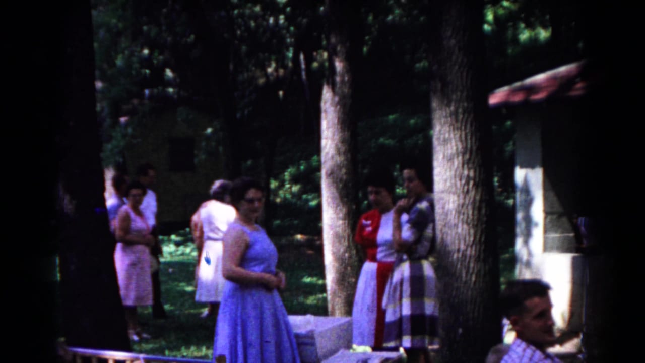 1963 Cold Springs home-movie frame showing women in blue, pink, red, and patterned dresses conversing among tall trees.
