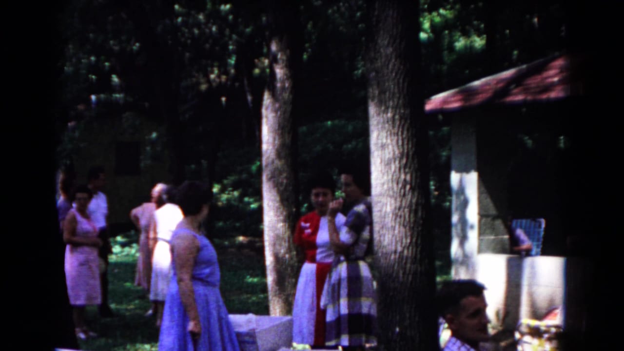 Women in dresses stand near trees and a small park structure.