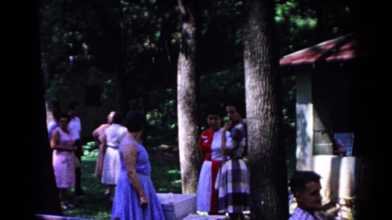 1963 Cold Springs park frame showing adults in mid-century dress grouped in conversation under heavy tree shade.