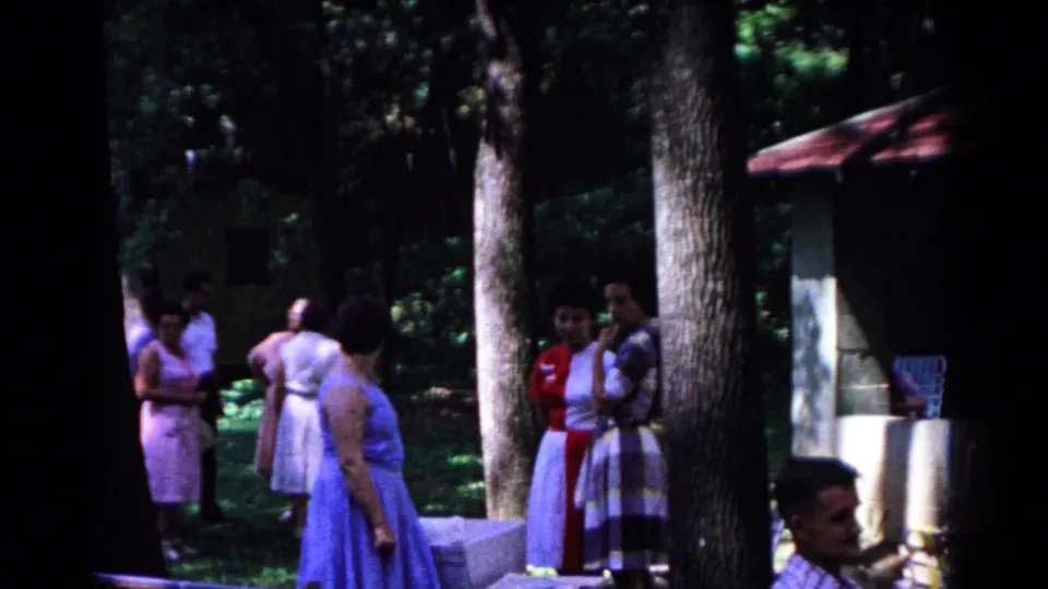 1963 Cold Springs park frame showing adults in mid-century dress grouped in conversation under heavy tree shade.