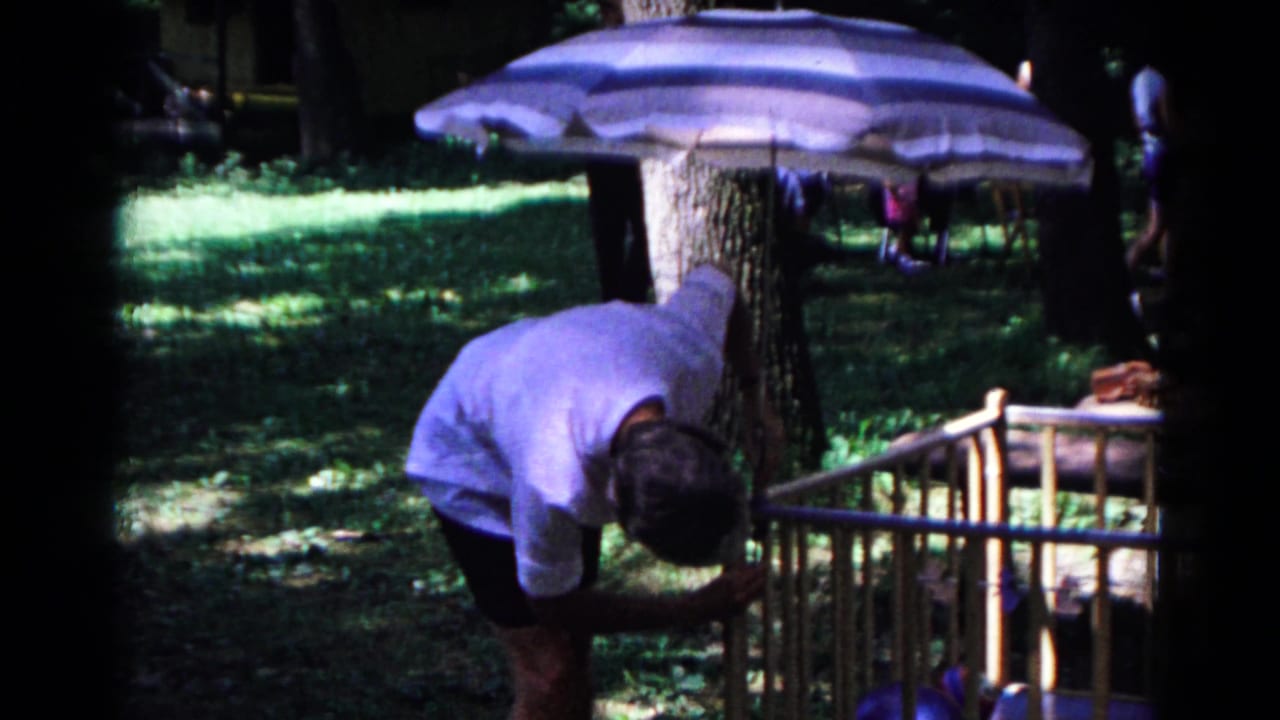 Cold Springs 1963 home-movie frame with a man bending over a wooden crib under a striped umbrella on shaded grass.