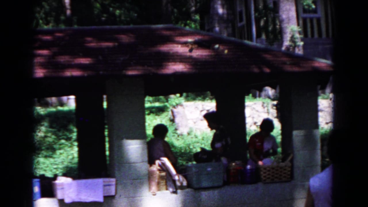 1963 Cold Springs home-movie frame showing adults seated under a red-roofed wooded pavilion with picnic supplies in shade.