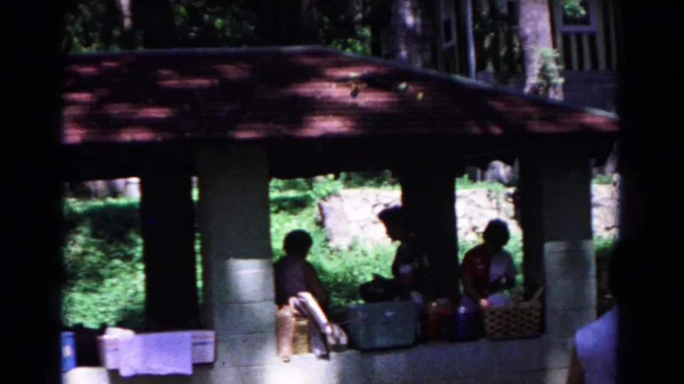 1963 Cold Springs home-movie frame showing adults seated under a red-roofed wooded pavilion with picnic supplies in shade.