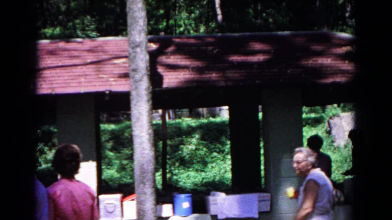 1963 Cold Springs frame showing adults moving near the campground pavilion, with picnic containers and wooded background.
