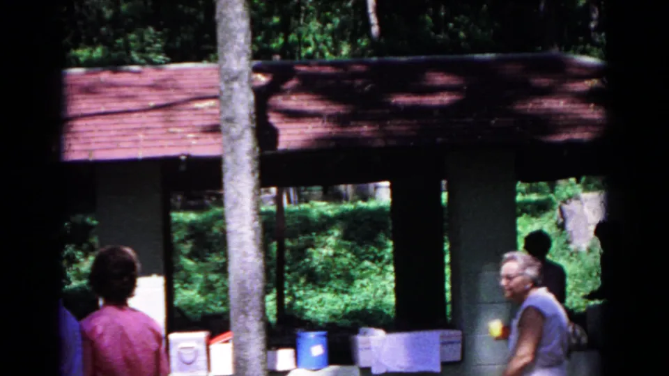1963 Cold Springs frame showing adults moving near the campground pavilion, with picnic containers and wooded background.