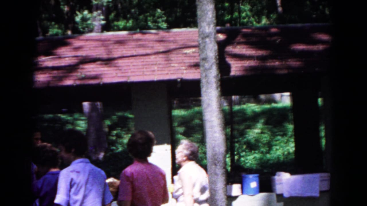 Cold Springs 1963 home-movie frame with adults in casual clothes gathered near coolers at a shaded campground shelter.