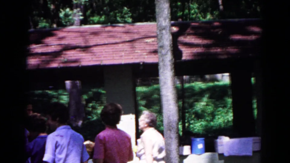 Cold Springs 1963 home-movie frame with adults in casual clothes gathered near coolers at a shaded campground shelter.