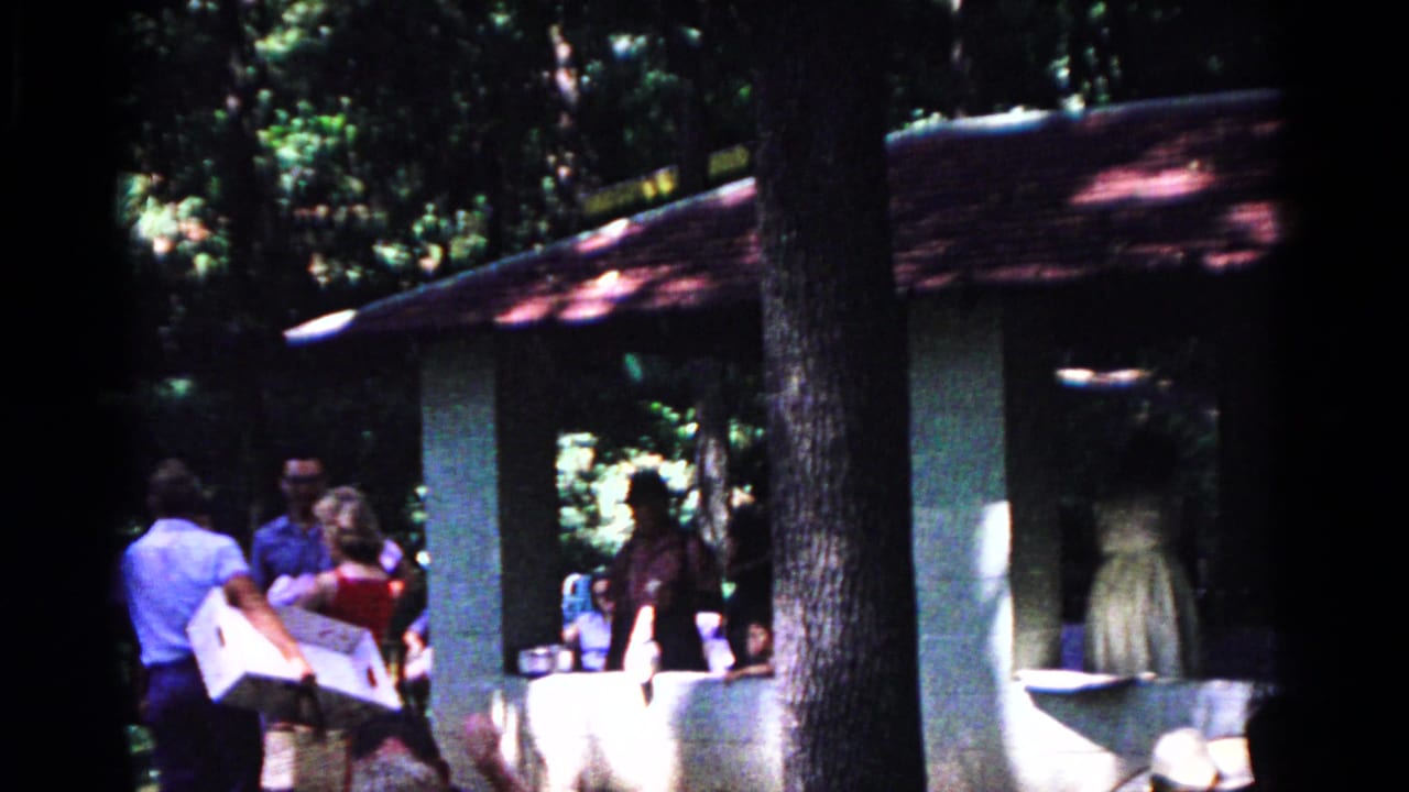 A man carries a large white item as adults gather near the pavilion in 1963 Cold Springs park footage.