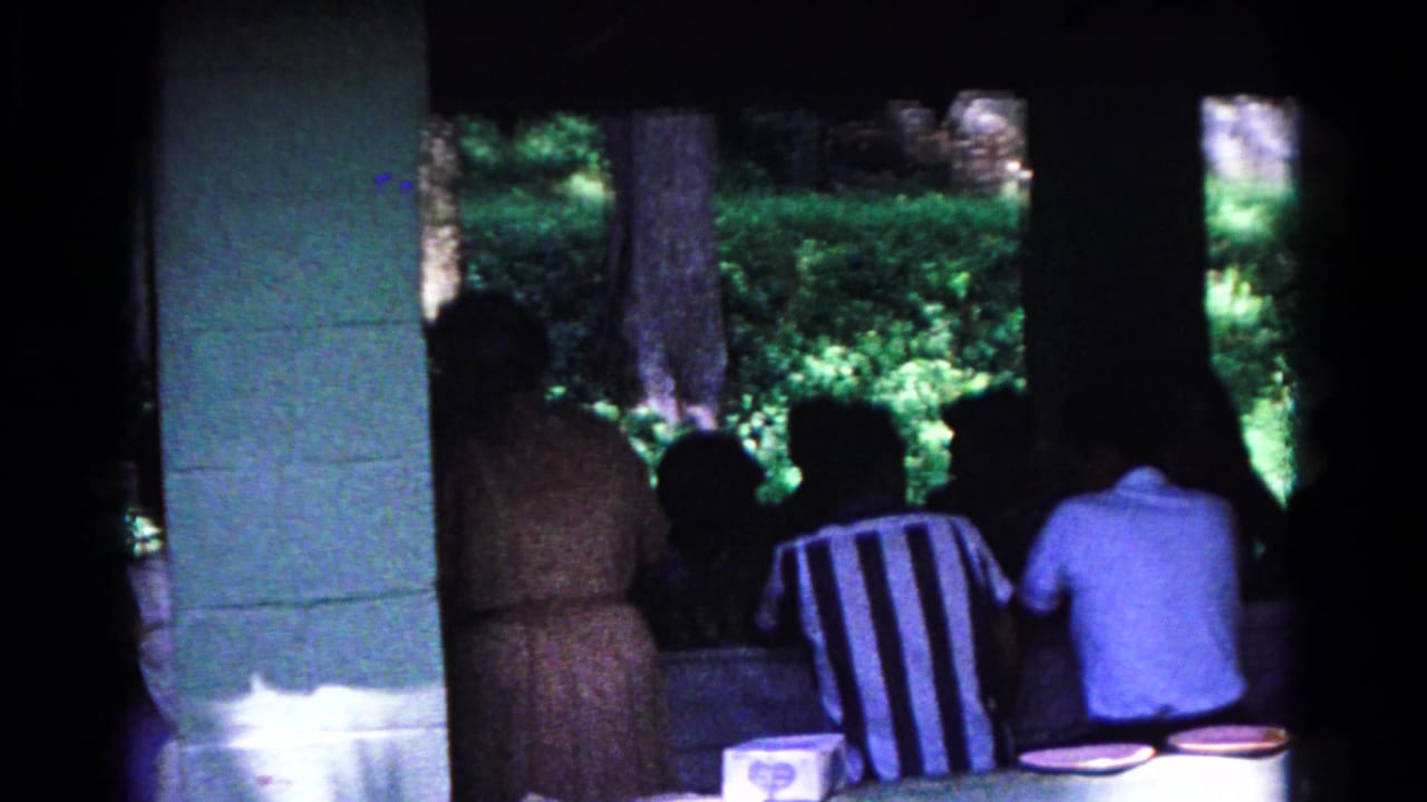 People seated at picnic tables inside a shaded pavilion with trees outside.