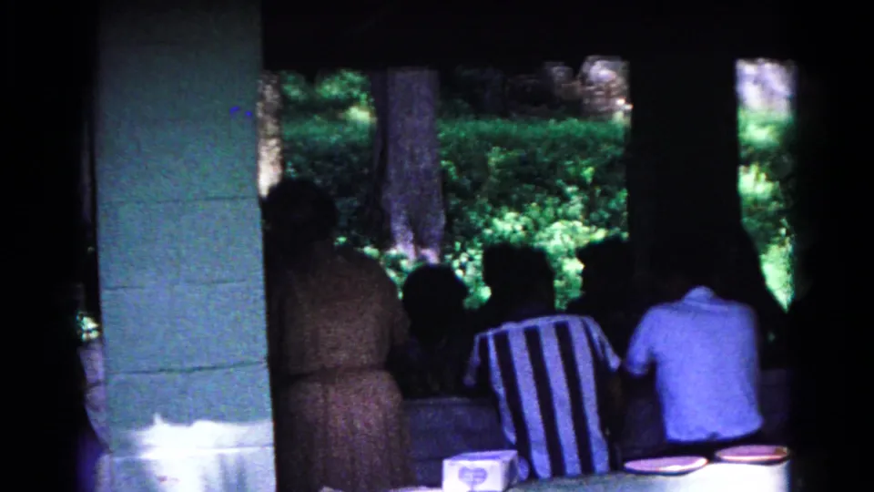 1963 Cold Springs home-movie frame showing people at picnic tables inside a shaded pavilion, with green trees beyond the open wall.