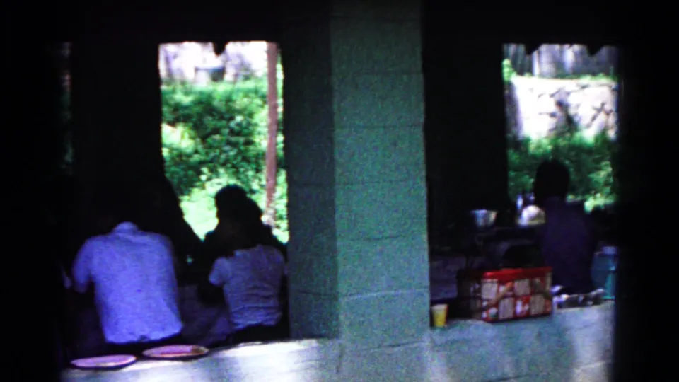 The final frame shows people seated along the pavilion table, with a block column, plates, a box or cooler, and bright greenery outside.