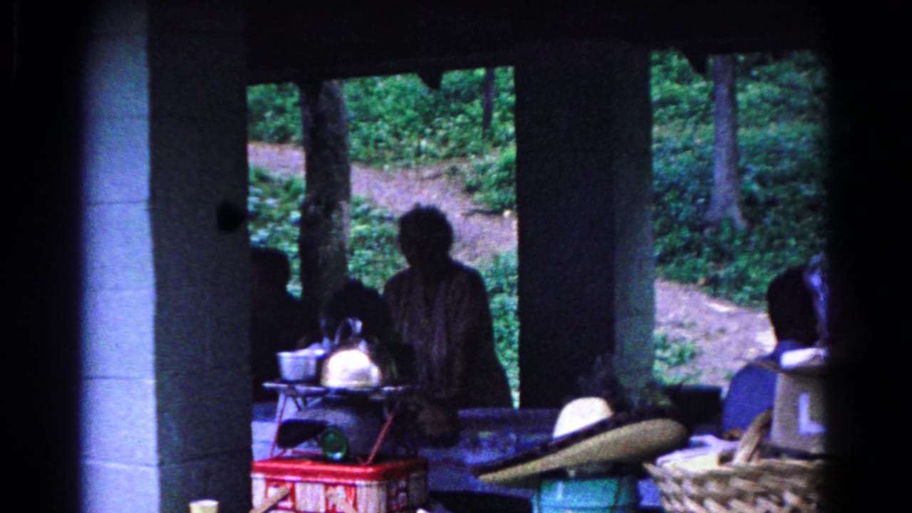 1963 Cold Springs home-movie frame showing people in shadow beneath a pavilion, with a wooded path and picnic items visible behind them.