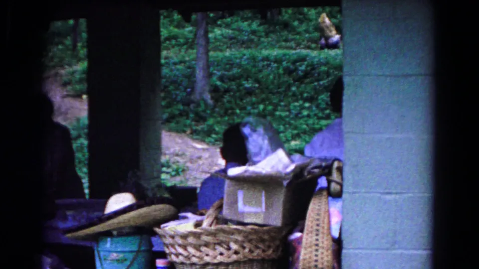 A closer 1963 home-movie view of picnic belongings under a pavilion, including baskets, containers, a hat, and woods outside.