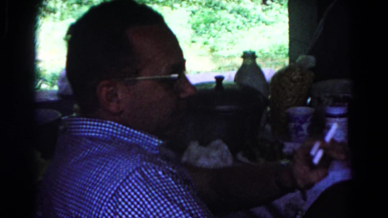 In 1963 Cold Springs home-movie footage, a seated man in glasses and a checkered shirt holds a cigarette near mugs and table items.