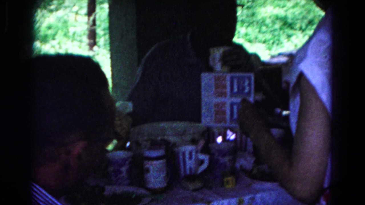 Adults sit around a dim table with mugs, cigarettes, and a visible HOT BUNS box.